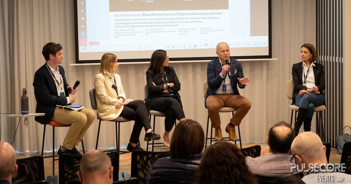 Five panellists seated on stools on a stage in front of a presentation screen, facing a seated audience